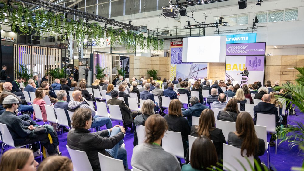 BAU 2025 visitors sitting on chairs, looking at a podium where a person is giving a presentation.