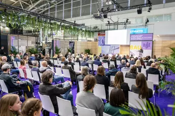 Auf Stühlen sitzende Besucher der BAU 2025. Sie blicken auf ein Podium, auf dem eine Person einen Vortrag hält.