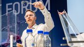 Ein Mann mit einer Laborschutzbrille steht an einem Tisch, auf dem Chemikalien in Flaschen stehen und hält etwas mit einer Zange in die Luft.