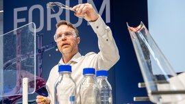 Ein Mann mit einer Laborschutzbrille steht an einem Tisch, auf dem Chemikalien in Flaschen stehen und hält etwas mit einer Zange in die Luft.