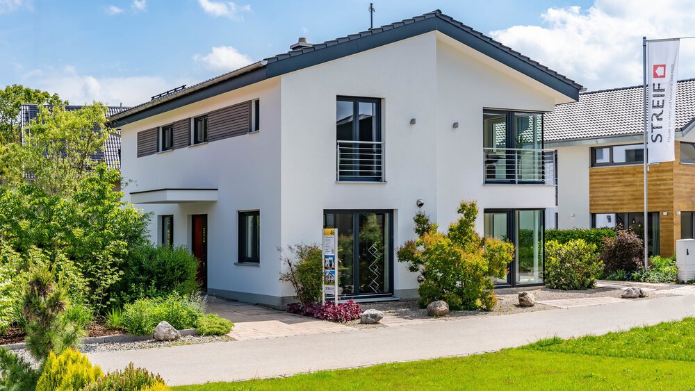 Ein modernes, zweistöckiges weißes Haus mit großen Fenstern, einem Balkon und einem gepflegten Vorgarten unter blauem Himmel. In der Nähe des Eingangs sind ein Schild und Grünpflanzen zu sehen.