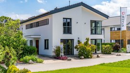 Ein modernes, zweistöckiges weißes Haus mit großen Fenstern, einem Balkon und einem gepflegten Vorgarten unter blauem Himmel. In der Nähe des Eingangs sind ein Schild und Grünpflanzen zu sehen.