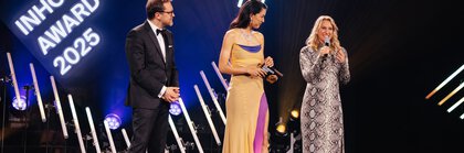 Three people in festive attire stand on the stage of the INHORGENTA AWARD 2025 and address the audience.