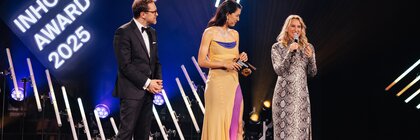 Three people in festive attire stand on the stage of the INHORGENTA AWARD 2025 and address the audience.