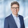 Ein Mann mit dunkler Brille und lockigen kurzen Haaren in einem blauem Anzug posiert lächelnd.