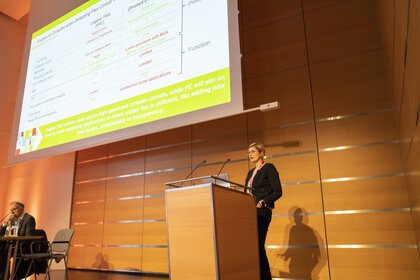 Eine Frau mit schwarzem Anzug und kurzen Haaren steht an einem Podium und hält in einem Konferenzraum eine Präsentation.