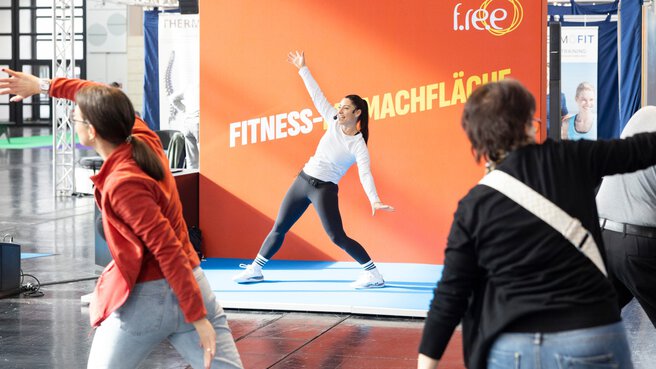 A trainer demonstrates an exercise in a fitness area in the exhibition hall, while several participants copy the movement. An orange wall with the words “Fitness-Mitmachfläche” (fitness activity area) can be seen in the background.
