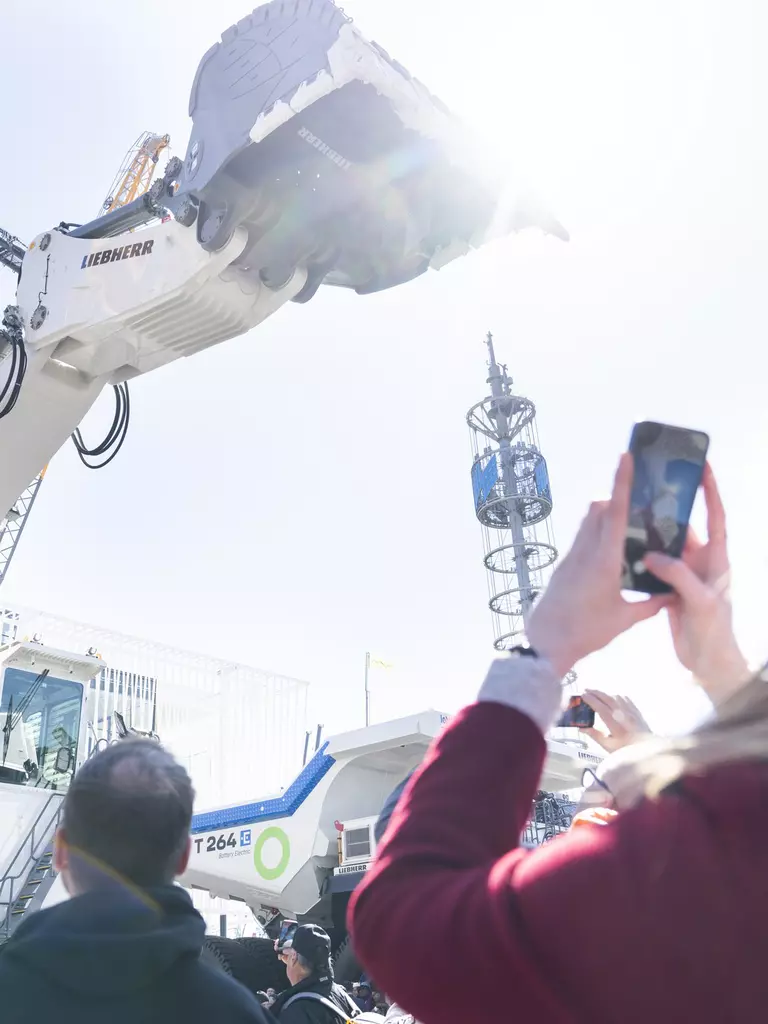 A woman takes a photo of a large white excavator stretching its shovel towards the sky.