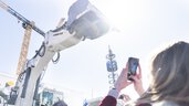 A woman takes a photo of a large white excavator stretching its shovel towards the sky.