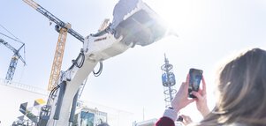 Eine Frau fotografiert einen großen weißen Bagger, der seine Schaufel gen Himmel streckt.