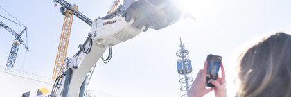 Eine Frau fotografiert einen großen weißen Bagger, der seine Schaufel gen Himmel streckt.
