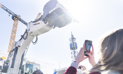 A woman takes a photo of a large white excavator stretching its shovel towards the sky.
