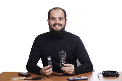 Ein Mann mit dunklen Haaren, Bart und schwarzem Pullover sitzt hinter einem Tisch, lächelt und hält technische Geräte in der Hand.
