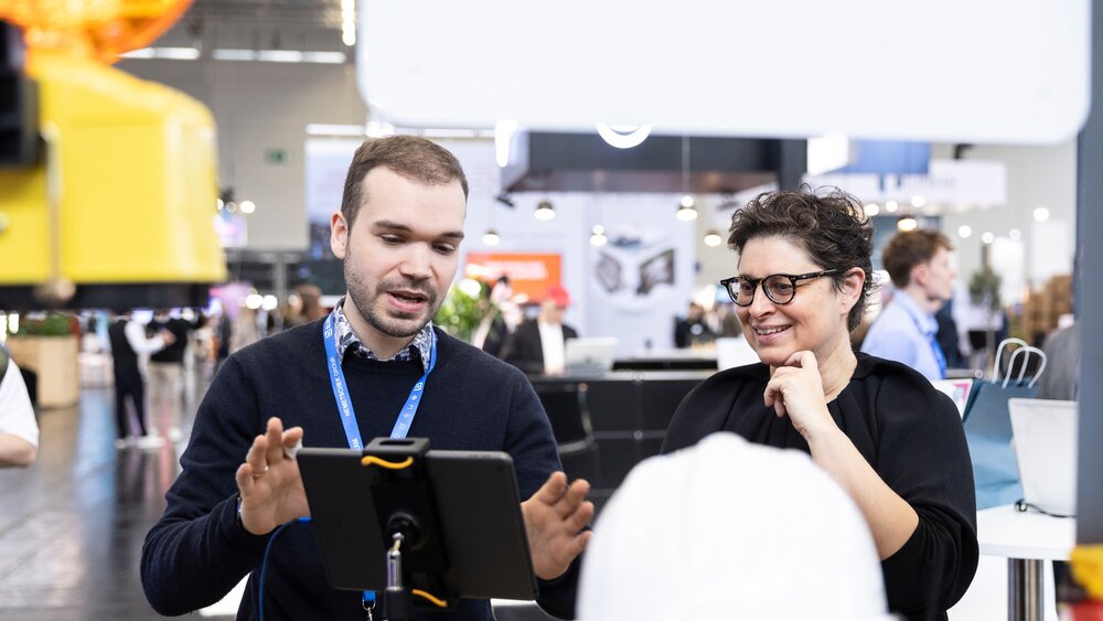 Ein Mann und deine Frau stehen an einem Messestand, sind im Dialog und blicken gemeinsam auf einen Screen.
