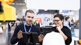 Ein Mann und deine Frau stehen an einem Messestand, sind im Dialog und blicken gemeinsam auf einen Screen.