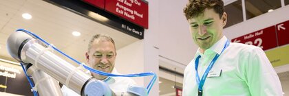 Zwei Männer an einer Messestation beobachten einen kollaborativen Roboterarm, der ein Bauteil greift – moderne Photonik-Innovation live auf der Messe.