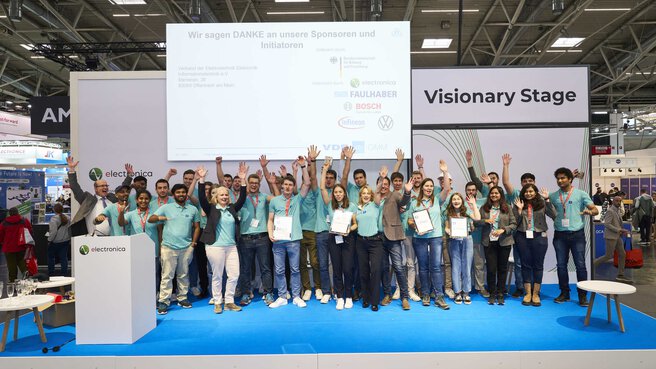 A group of people in turquoise shirts stand cheering on the stage of the electronica Visionary Stage.