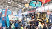 Several visitors walk through a large exhibition hall with stands showcasing water sports, surfing, and kite surfing equipment. Large advertising banners for the brands hang above the exhibition areas.