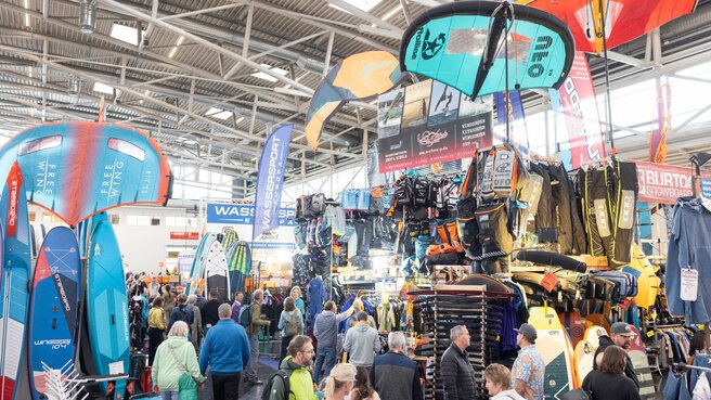 Several visitors walk through a large exhibition hall with stands showcasing water sports, surfing, and kite surfing equipment. Large advertising banners for the brands hang above the exhibition areas.