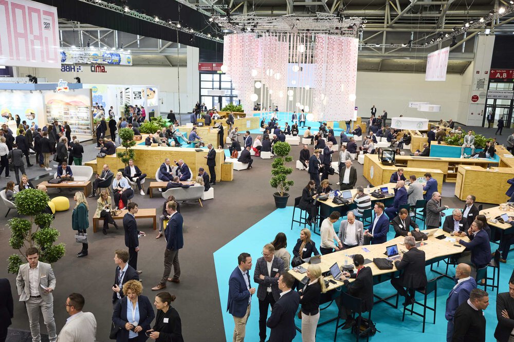 View of a spacious area in an exhibition hall with numerous seating options, tables and many visitors.
