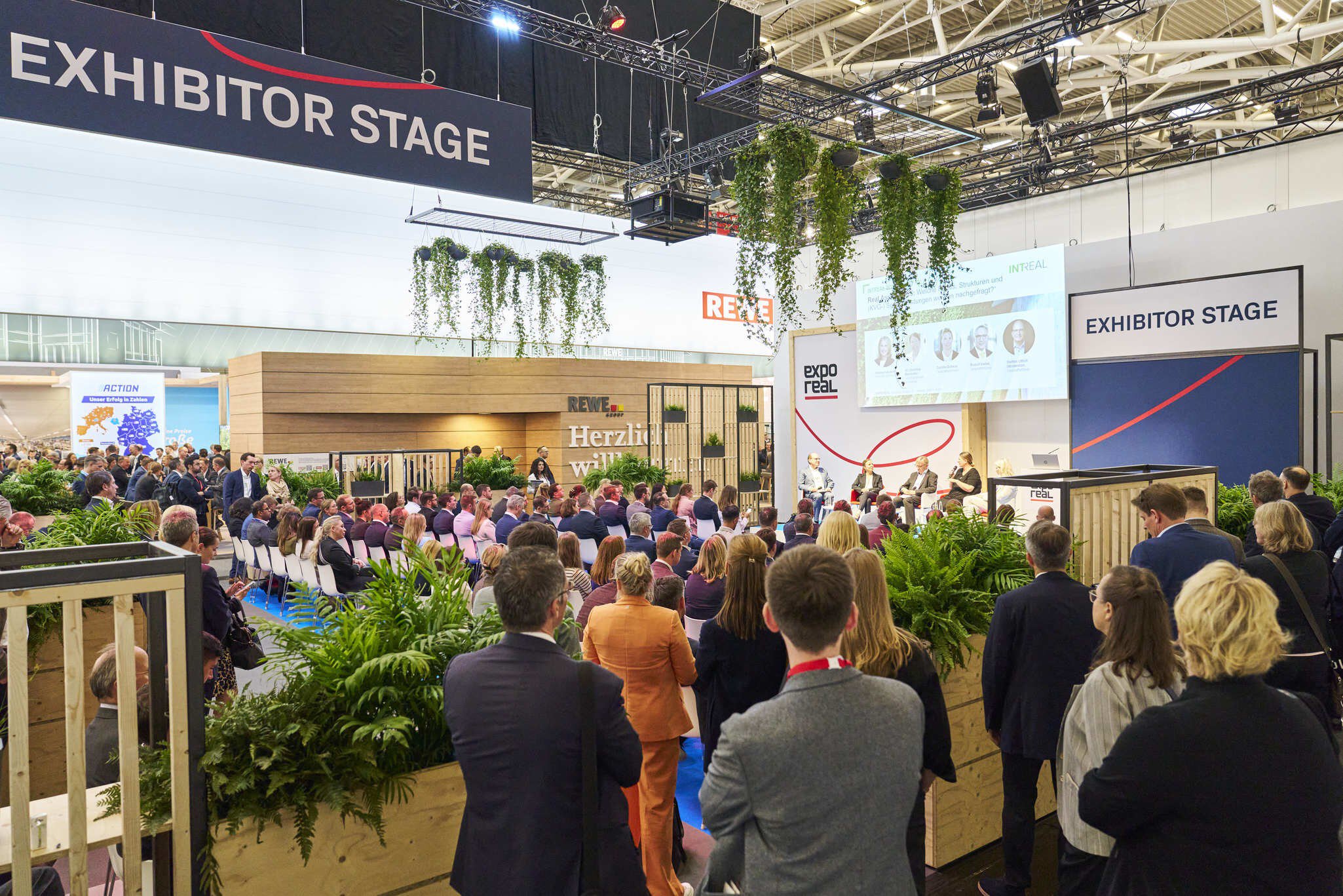 A crowd gathers around an exhibitor's booth at a trade fair featuring plants and banners in a large indoor space.