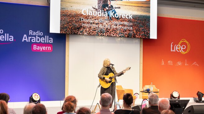A woman wearing a leopard-print top and holding a guitar stands on stage at f.re.e and sings into a microphone in front of an audience.