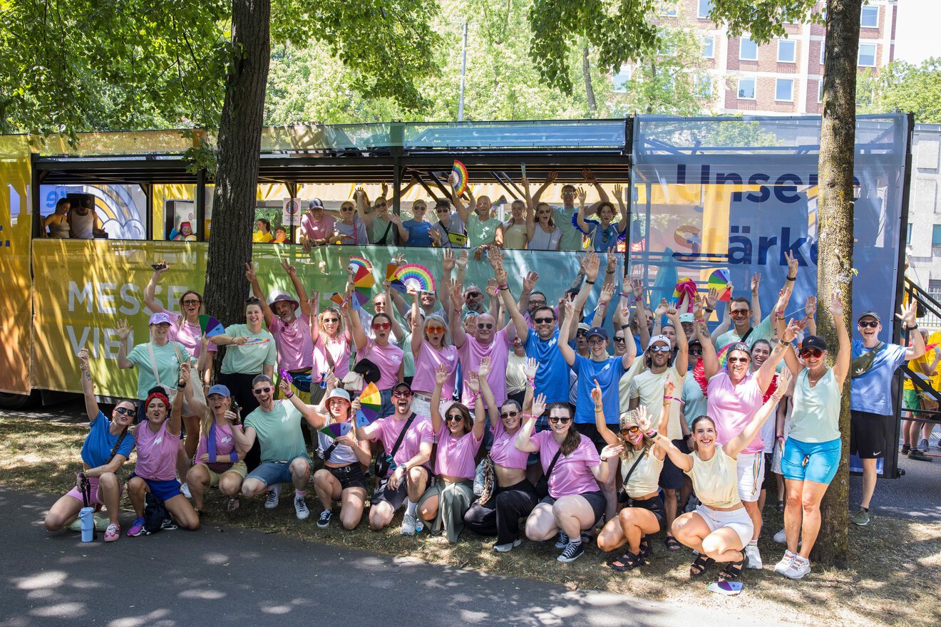 Eine große Gruppe von Menschen in bunten Hemden jubelt und posiert gemeinsam im Freien vor einem geschmückten Bus.
