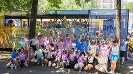 A large group of people wearing colorful shirts are cheering and posing together outdoors in front of a decorated bus.