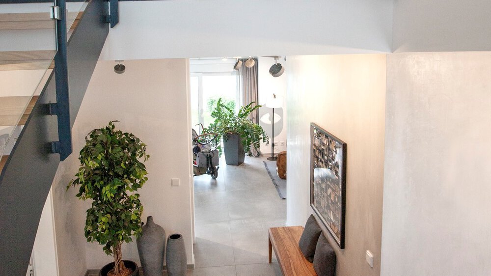 Modernes Interieur mit Treppen, großen Pflanzen und durch ein Fenster einfallendem Licht, das eine helle, luftige Atmosphäre schafft.