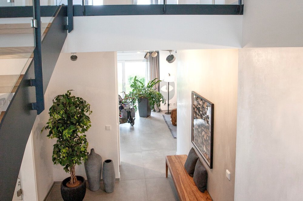 Modernes Interieur mit Treppen, großen Pflanzen und durch ein Fenster einfallendem Licht, das eine helle, luftige Atmosphäre schafft.