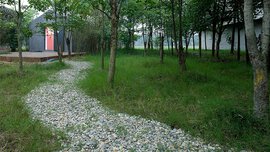 Ein gewundener Schotterweg führt durch eine Rasenfläche mit Bäumen zu einem kleinen Gebäude mit einer rosa Tür.