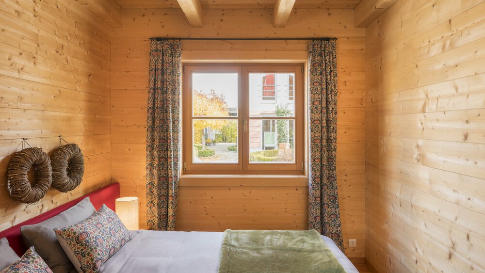 Gemütliches Zimmer mit einem Bett, einer grünen Decke, Kissen und einem Fenster mit gemusterten Vorhängen, Holzwänden und Deckenbalken.