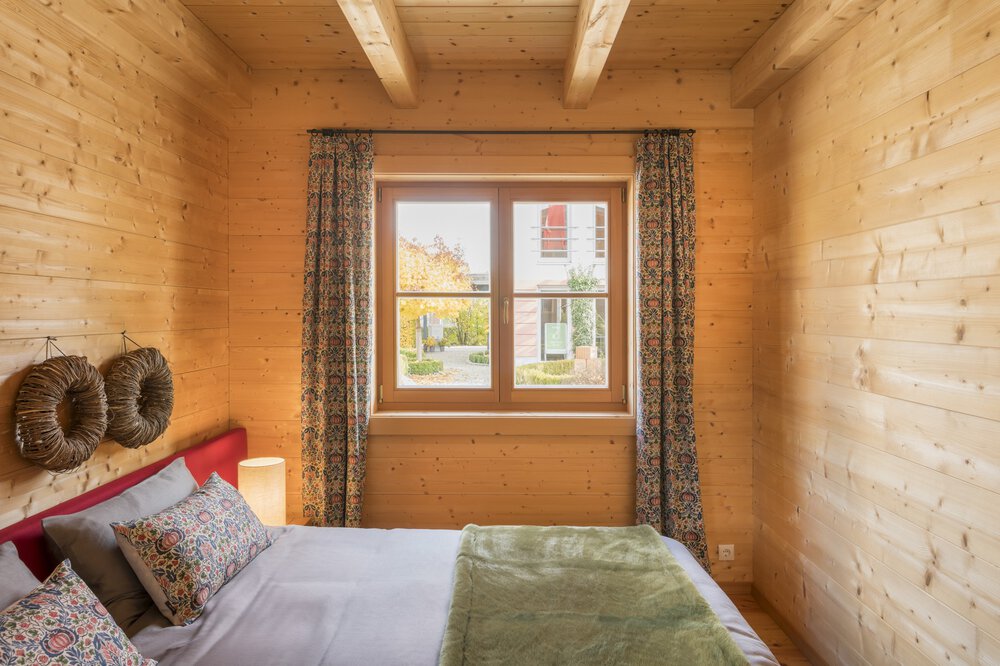 Gemütliches Zimmer mit einem Bett, einer grünen Decke, Kissen und einem Fenster mit gemusterten Vorhängen, Holzwänden und Deckenbalken.