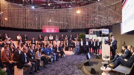Ein Vortrag auf der EXPO REAL in München mit zahlreichen Zuhörerinnen und Zuhörern, die auf gestuften Sitzplätzen sitzen und aufmerksam dem Sprecher auf der Bühne folgen. Der Referent steht an einem Rednerpult, während neben ihm weitere Diskussionsteilnehmer auf Sesseln Platz genommen haben. Über der Szene hängen dekorative Seile, die eine moderne Atmosphäre schaffen, und im Hintergrund sind weitere Messestände zu erkennen.