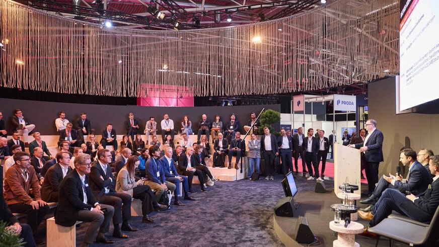 A presentation at EXPO REAL in Munich with numerous attendees seated on tiered benches, listening attentively to a speaker on stage. The presenter stands at a podium, while other panelists are seated beside him in chairs. Decorative hanging ropes above the area create a modern atmosphere, and additional exhibition booths can be seen in the background.