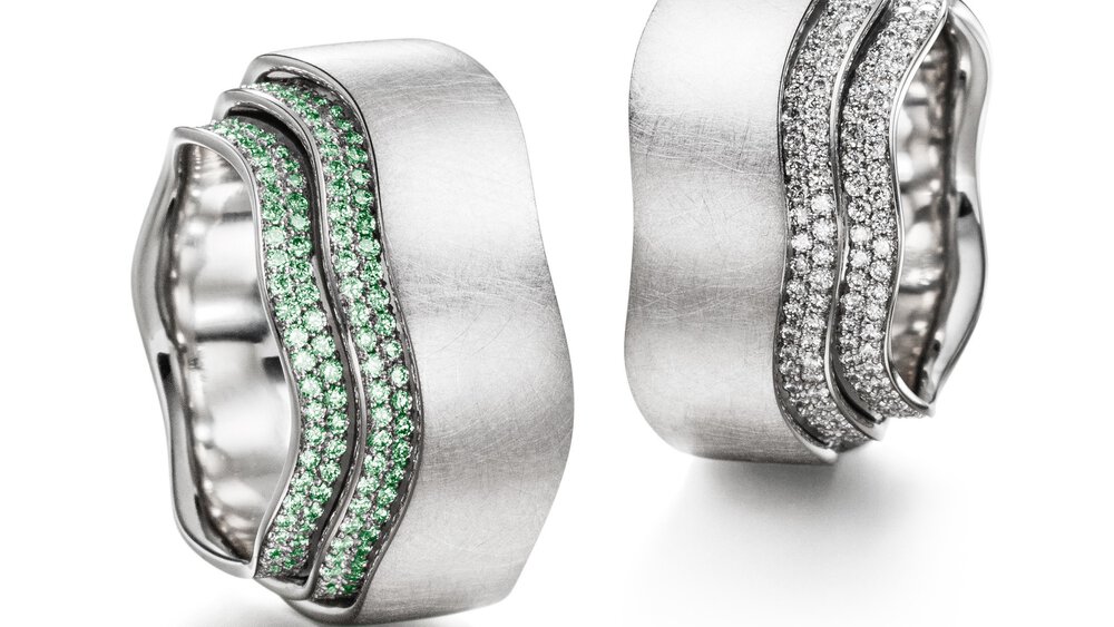 Two ornate, almost identical wavy rings made of silver and set with many small diamonds, one with green and the other with white.