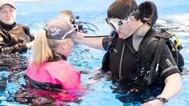 A woman explains diving equipment to a man in the swimming pool.