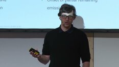 Ein Redner hält einen Vortrag auf der Bühne und bedient mit einer Fernbedienung die Textpräsentation im Hintergrund.