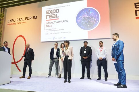 Sieben Personen stehen bei der Verleihung der Expo Real Forum 2024 Impact Awards auf der Bühne, ein Redner steht am Mikrofon.