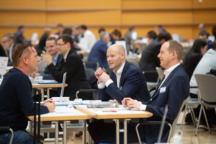 Three men in suits sitting at a table chatting; other groups in the background.