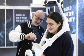 A woman is wearing an Exco skeleton with camera-device, and a man is explaining how to use the device.