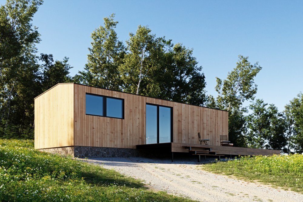 Moderne Holzhütte mit großen Fenstern, inmitten einer Grasfläche mit umgebenden Bäumen unter einem klaren blauen Himmel.