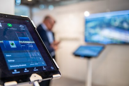 Tablet mit fokussierter Bedienoberfläche, im Hintergrund unscharf eine Person neben einem Bildschirm stehend.