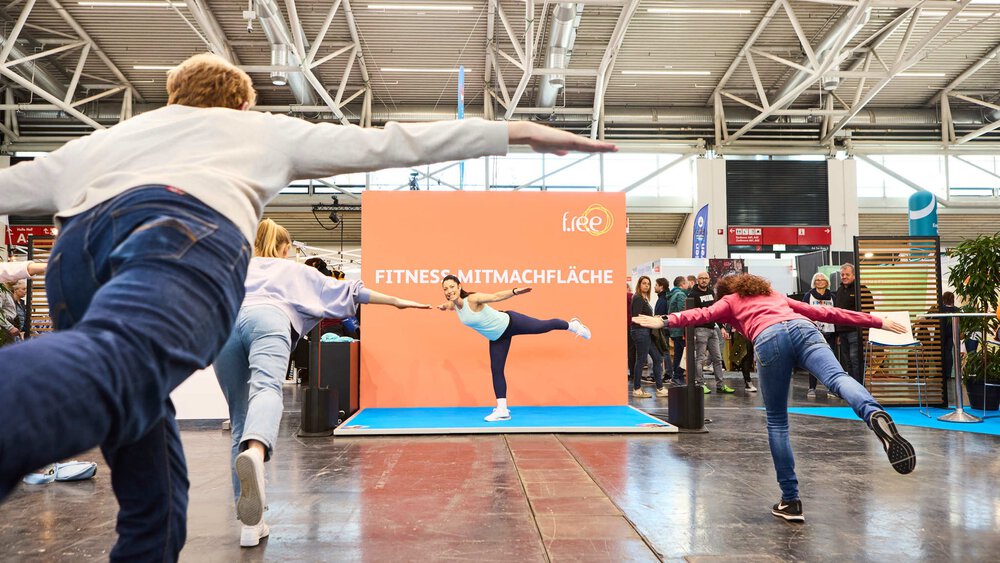Eine Frau in Sportkleidung steht mit ausgestreckten Armen auf einem Bein auf einer Bühne der f.re.e, während drei weitere Personen ihre Haltung nachahmen.
