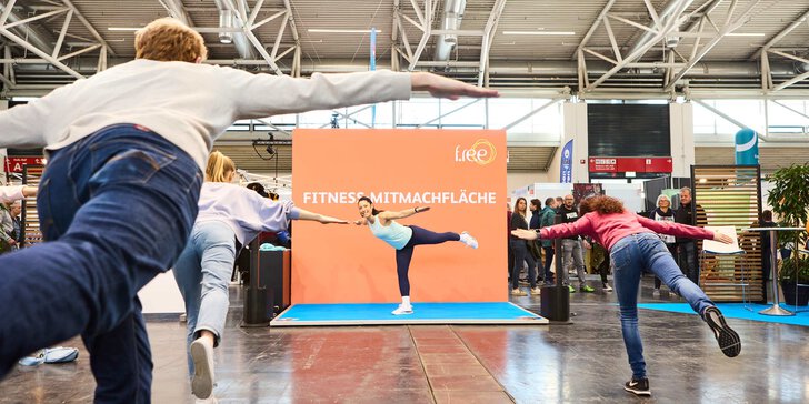 Eine Frau in Sportkleidung steht mit ausgestreckten Armen auf einem Bein auf einer Bühne der f.re.e, während drei weitere Personen ihre Haltung nachahmen.