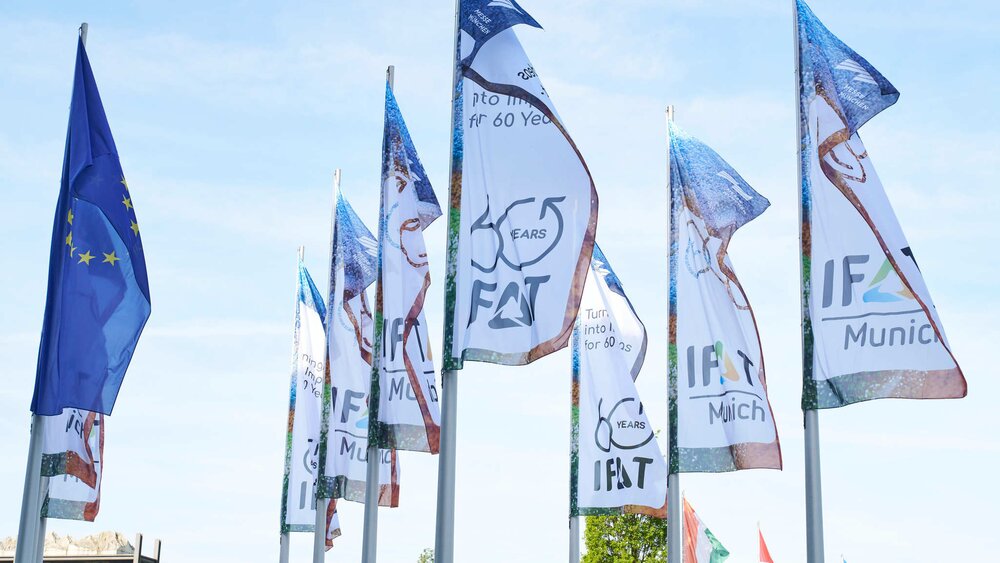 Row of flagpoles with waving blue EU flags and white IFAT trade fair flags against a lightly cloudy sky.