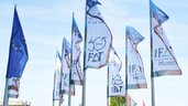 Reihe von Flaggenmasten mit wehenden blauen EU-Flaggen und weißen IFAT-Messeflaggen vor leicht bewölktem Himmel.