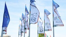 Row of flagpoles with waving blue EU flags and white IFAT trade fair flags against a lightly cloudy sky.