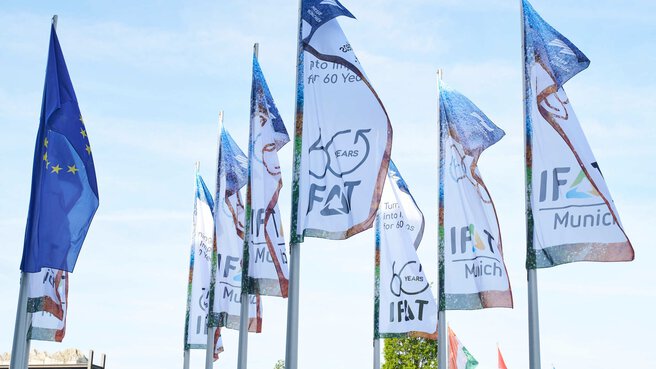 Reihe von Flaggenmasten mit wehenden blauen EU-Flaggen und weißen IFAT-Messeflaggen vor leicht bewölktem Himmel.
