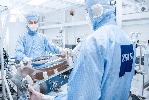 Two workers in cleanroom suits move a heavy frame in which an oval, curved mirror is fixed.
